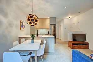 Une cuisine moderne avec un îlot central, des chaises de salle à manger blanches, un luminaire suspendu et un téléviseur à écran plat intégré dans un meuble de rangement en bois.