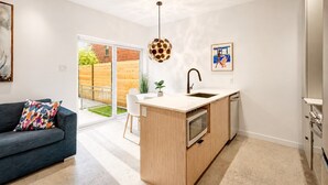 A modern kitchen with a central island, a built-in oven, and a sink. There is a dining area with a sofa and a colorful pillow. A large window provides a view of the outside. - Perfect 2bdr in the hart of Montreal (Montréal)