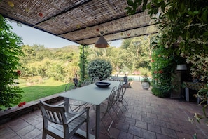 Un patio al aire libre con una mesa y sillas, rodeado de abundante vegetación y vistas a una piscina.