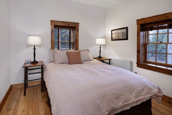 A bedroom with a large bed, two bedside tables, two lamps, a window, and a radiator. - Large Log Cabin in the Heart of the ADKS (Keene)