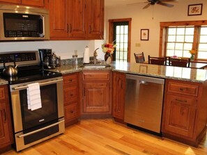 A kitchen with wooden cabinets, stainless steel appliances, a granite countertop, and a dining area with chairs. - Large Log Cabin in the Heart of the ADKS (Keene)