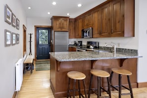 A kitchen with wooden cabinets, a stainless steel refrigerator, a microwave, and a granite countertop. There are three wooden stools at the island.