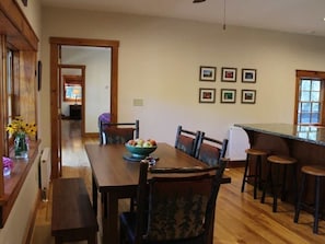 Een eethoek met een houten tafel, stoelen en een schaal fruit. Er hangen afbeeldingen in lijsten aan de muur en een raam met uitzicht.