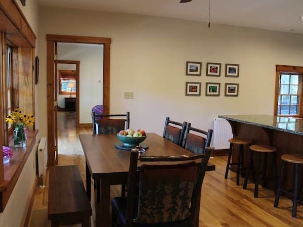 A dining area with a wooden table, chairs, and a bowl of fruit. There are framed pictures on the wall and a window with a view.