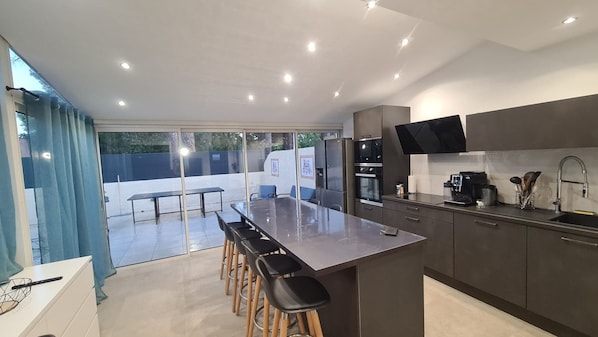 Cozinha moderna com ilha central, banquetas, forno embutido, micro-ondas, máquina de café e uma grande janela com vista para uma área externa cercada.