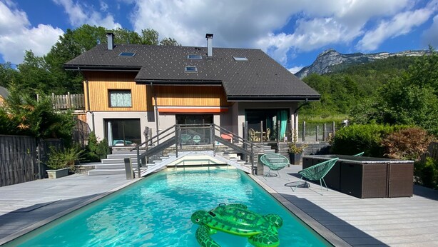 A modern house with a swimming pool, a wooden deck, and a mountain backdrop.