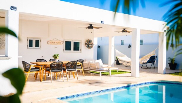 Zona junto a la piscina con una mesa y sillas, una hamaca y un letrero que dice "BEACH HOUSE".