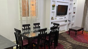 Uma área de refeições com mesa de vidro e cadeiras, um tapete estampado e uma televisão numa parede branca.