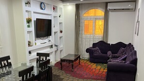Uma sala de estar com uma mesa de jantar de vidro, cadeiras de madeira escura, um sofá roxo e um tapete colorido com padrões.