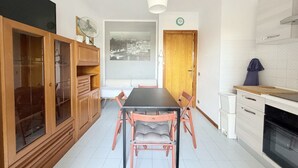 Zona pranzo con tavolo e sedie, un mobile contenitore in legno con ante in vetro, una ventola a soffitto bianca, un quadro con vista sulla città e una zona cucina con fornello e pensili.