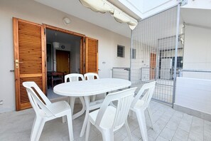 Un patio exterior con una mesa redonda y sillas blancas de plástico, puertas de madera y una reja de metal.