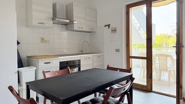 Una cucina moderna con mobili bianchi, un tavolo da pranzo nero e una porta scorrevole in vetro che dà su uno spazio esterno.