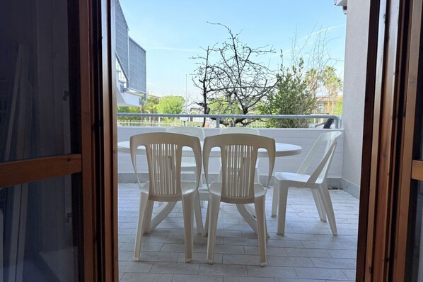 Un balcón con sillas de plástico y una mesa, una valla blanca y un árbol al fondo.