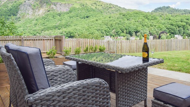 A patio with wicker furniture, a table with a bottle of champagne, and a view of a green hillside. - Craigmore View-UK34220 (Aberfoyle)