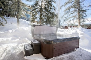 Un bain à remous installé à l’extérieur, dans un décor enneigé, avec des arbres et un bâtiment en arrière-plan.