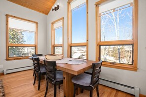 Un coin repas avec une table en bois et quatre chaises, de grandes fenêtres donnant sur des arbres et des bâtiments, et un plafond avec des poutres en bois.