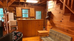 Interior acolhedor de uma cabana, com um banco de madeira, uma cadeira preta, uma pequena mesa e uma área de cozinha com armários de madeira e uma geladeira.
