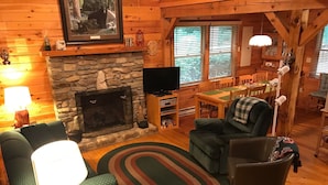 Interior acolhedor de uma cabana, com lareira de pedra, paredes de madeira, televisão e área de jantar.