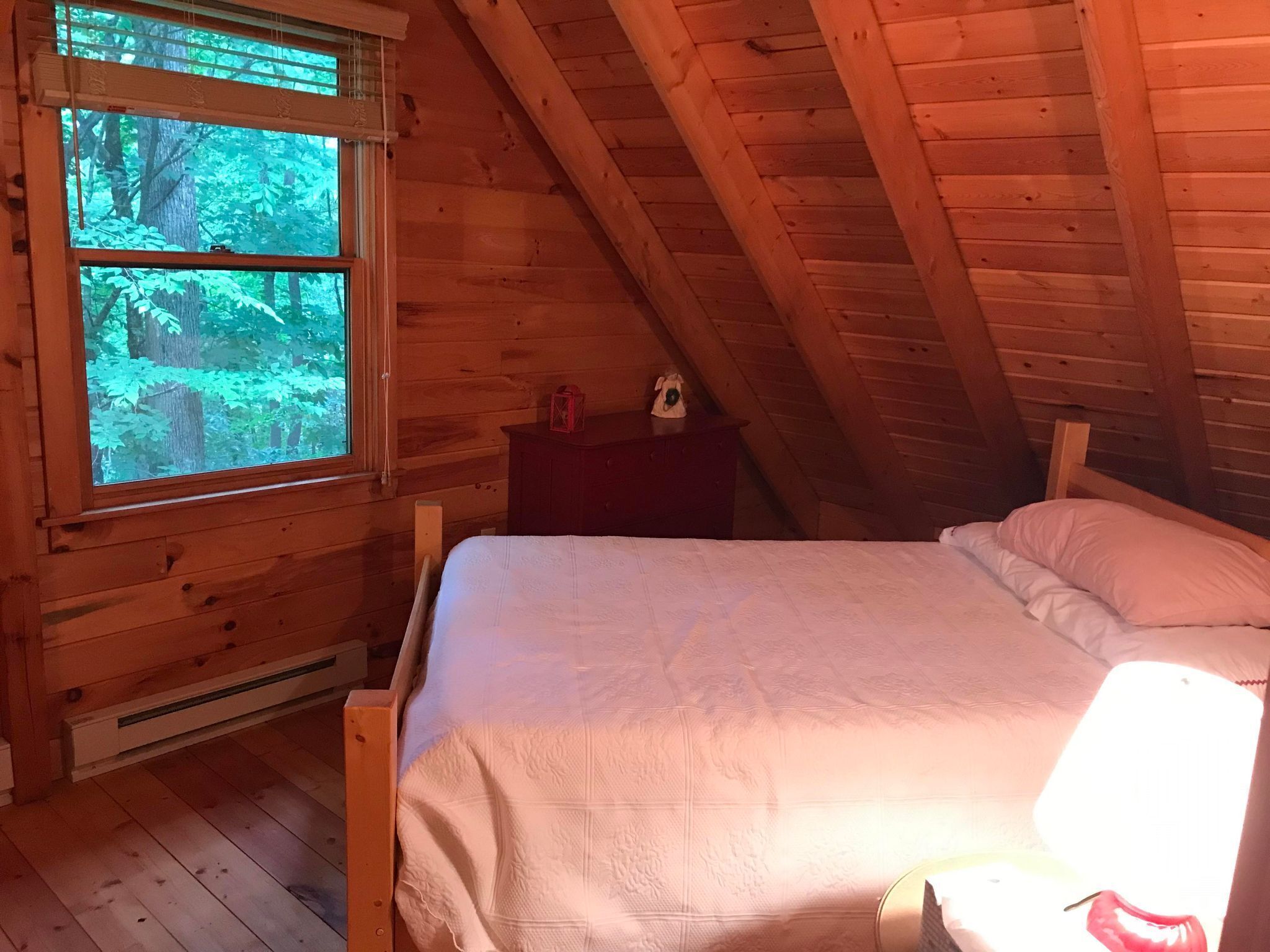 Un dormitori de cabana de fusta amb un llit, una tauleta de nit i una finestra amb vista a un bosc.