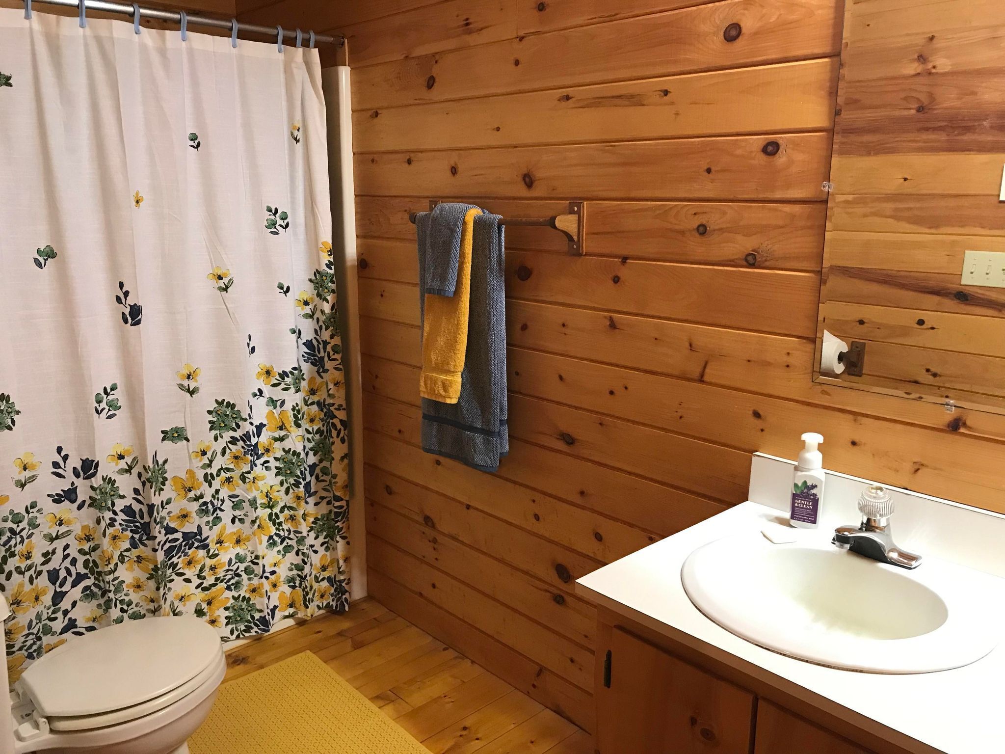 A bathroom with wooden walls, a toilet, a sink with a mirror, and a shower curtain with floral patterns.