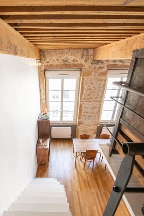 A narrow staircase leading to a dining area with a wooden table and chairs, a window, and a stone wall.