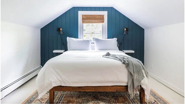 A bedroom with a sloped ceiling, a bed with white bedding, two bedside lamps, and a window with blinds. - Modern Mountain Getaway with Fire Pit (Shandaken)