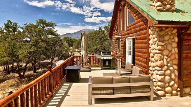 A log cabin with a stone chimney, a deck with outdoor furniture, and a view of trees and mountains. - The Stargazer-Your private retreat in the mountains! (Crestone)