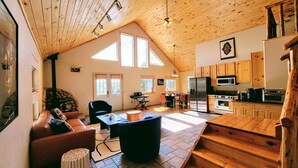 A spacious living area with a high ceiling, wooden beams, a fireplace, a dining table, and a kitchen with modern appliances. - The Stargazer-Your private retreat in the mountains! (Crestone)
