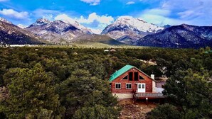 A cabin with a green roof and a wooden deck, surrounded by dense forest and mountains in the background. - The Stargazer-Your private retreat in the mountains! (Crestone)