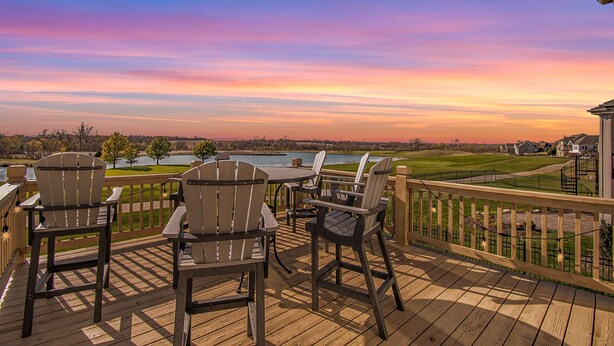Une terrasse en bois avec des fauteuils Adirondack et une table, offrant une vue sur un lac et un terrain de golf au coucher du soleil.