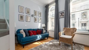 Una sala de estar moderna con un sofá azul, un sillón de mimbre y un ventanal con cortinas.
