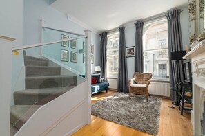 Eine Treppe mit Glasgeländern führt zu einem Wohnbereich mit blauem Sofa, Korbsessel und Kamin.