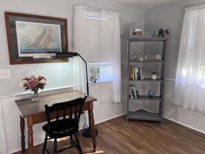 A room with a wooden desk, a chair, a bookshelf, a framed painting, and a window with curtains.