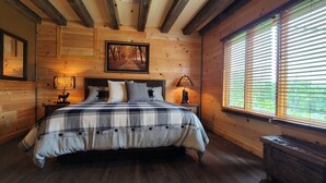 A cozy bedroom with a wooden floor, a large bed with a plaid bedspread, two bedside lamps, a framed picture on the wall, and a window with blinds. - Beautiful Home with Lake Views 4/2, Beautiful Home with Lake Views 4/2 - Resort Access for Fee (Lake George)