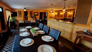 A dining area with a long table set for six, wooden walls, and a kitchen area with a bar. - Beautiful Home with Lake Views 4/2, Beautiful Home with Lake Views 4/2 - Resort Access for Fee (Lake George)