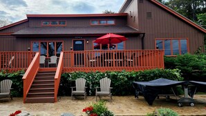 A two-story building with a red exterior, a covered patio area with chairs, and a red umbrella. - Beautiful Home with Lake Views 4/2, Beautiful Home with Lake Views 4/2 - Resort Access for Fee (Lake George)