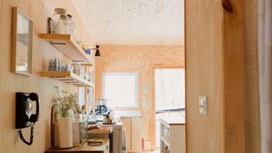 A wooden interior with a kitchen area, wooden shelves with jars, a telephone, and a window providing natural light.