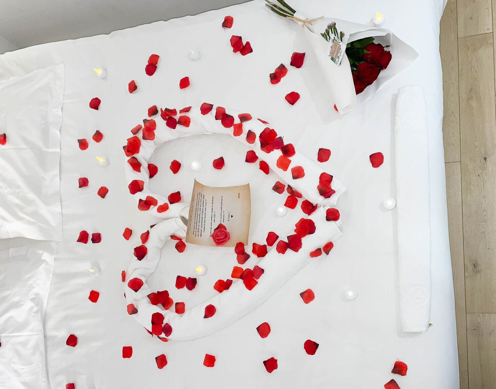Un lit blanc orné de pétales de roses disposés en forme de cœur, une bougie et un bouquet de roses rouges.