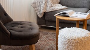 Een gezellige woonkamer met een bruine fauteuil, een grijze bank, een klein houten bijzettafeltje met een open boek, en een zachte witte poef.