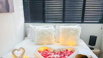 A bed with white bedding, two fluffy pillows, a heart-shaped decoration with candles, and a bouquet of red roses.