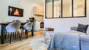 Una sala de estar moderna con un sofá, un comedor con sillas, una cocina con armarios y un televisor mostrando un fuego.
