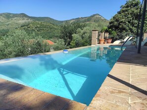 A rectangular pool with clear blue water, surrounded by a tiled deck. In the background, there are green hills and a clear sky.