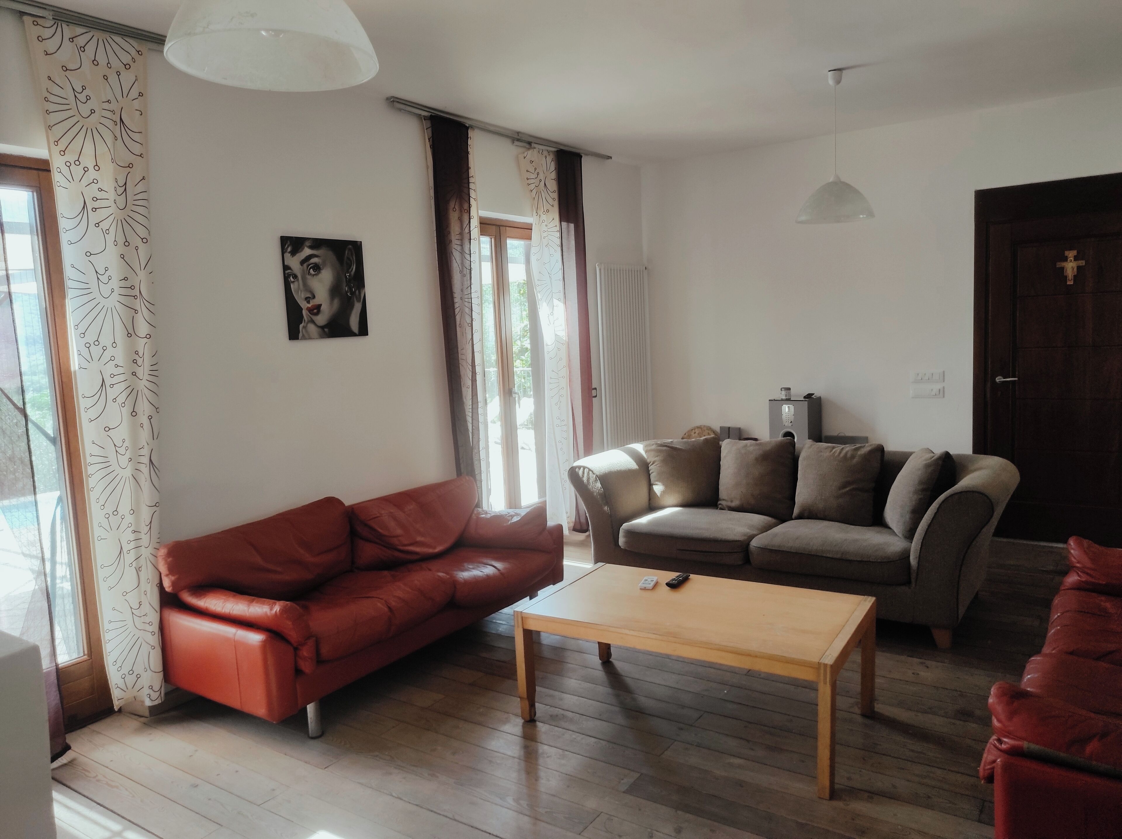 A living room with a red leather sofa, a beige sofa with multiple cushions, and a wooden coffee table. There is a painting on the wall, a ceiling light, and a door with a cross.