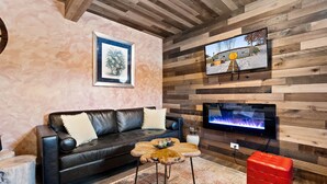 A modern living room with a black leather sofa, a wooden accent wall, a flat-screen TV mounted above a fireplace, and a red ottoman. - Rustic Hobbit Inspired Hocking Hills State Park 3 mi away Hot Tub Pet Friendly  (Creola)