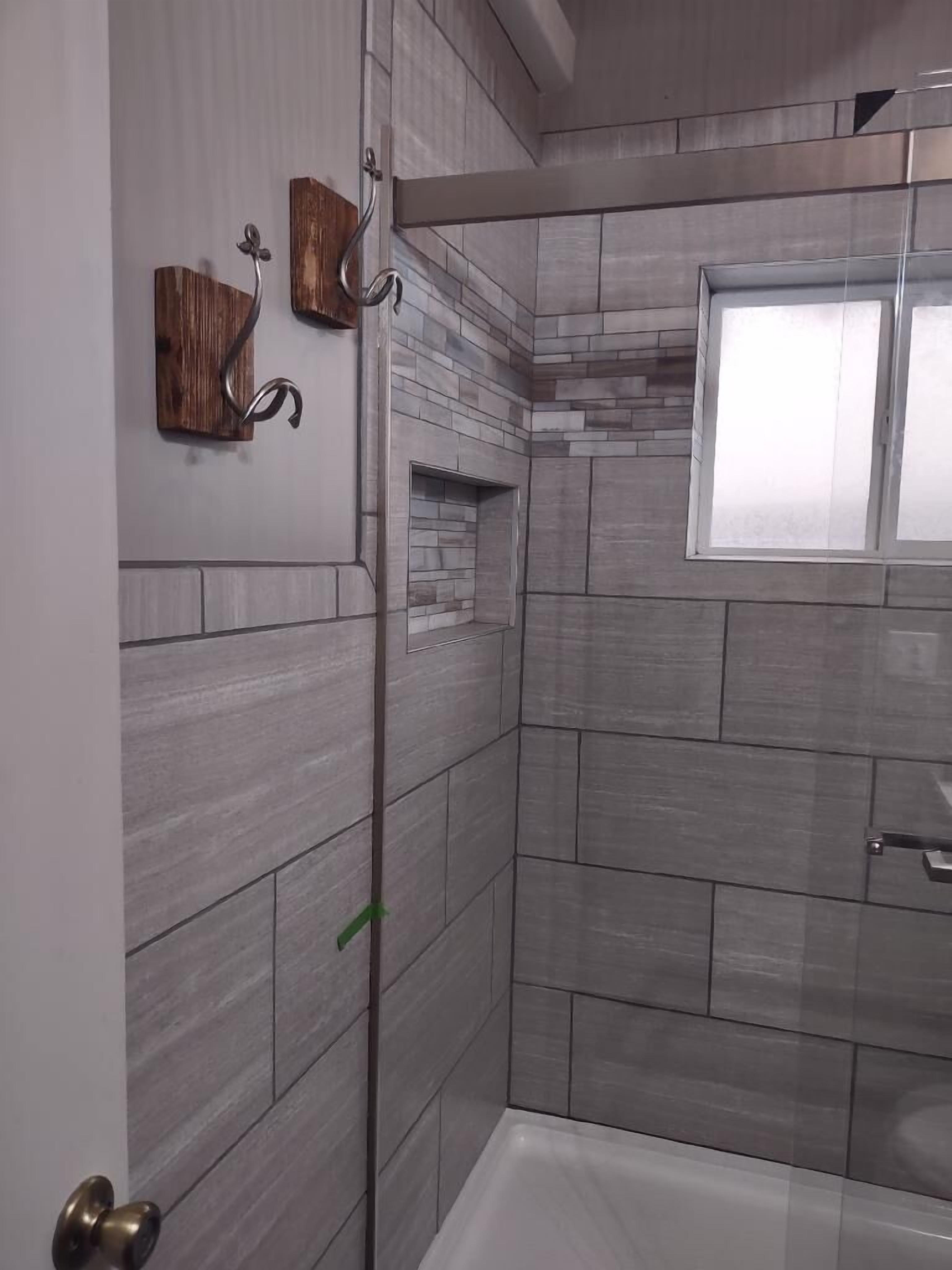 A modern bathroom with grey tiles, a glass shower enclosure, a wooden accent wall, and a window.