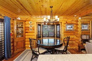 Interior de una cabaña de madera con una mesa redonda para comer rodeada de sillas, un mueble de madera con puertas de cristal y un candelabro.