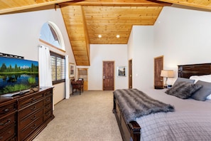 Un dormitorio con techo de madera, una cama grande con una manta de piel, un televisor sobre un aparador y una puerta que da a otra habitación.