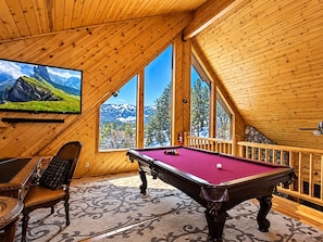 Una habitación de madera con una mesa de billar, un sillón cómodo y un televisor que muestra un paisaje montañoso.
