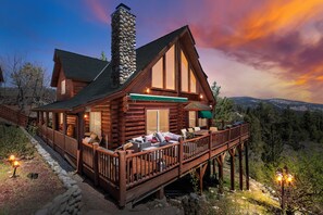 A log cabin with a deck and outdoor seating area, surrounded by a stone wall and a scenic view of mountains.