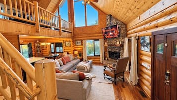 Spazioso interno di una baita di legno con soffitto alto, camino, zona pranzo e balcone.